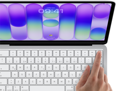 MacBook Neo, gümüş rengi, ekranda oturum açma penceresi gösteriliyor, kullanıcının parmağı klavyenin sağ üst köşesindeki Touch ID sensörüne dokunuyor