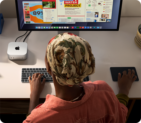 Masada oturan bir kişi Mac mini ile yazı yazıyor, Studio Display bağlı, Magic Keyboard, Magic Trackpad ve çeşitli eşyalar