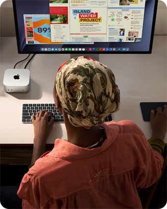 Masada oturan bir kişi Mac mini ile yazı yazıyor, Studio Display bağlı, Magic Keyboard, Magic Trackpad ve çeşitli eşyalar