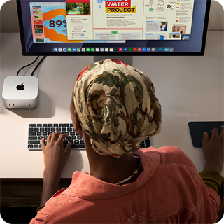 Masada oturan bir kişi Mac mini ile yazı yazıyor, Studio Display bağlı, Magic Keyboard, Magic Trackpad ve çeşitli eşyalar