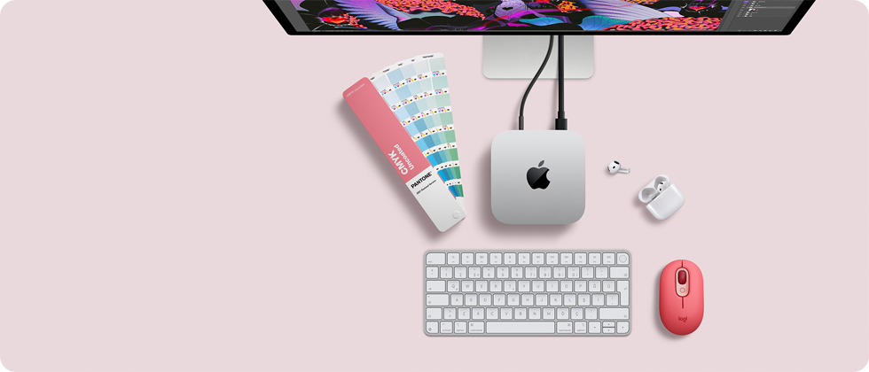 Mac mini, üstten görünüm, gümüş rengi alüminyum, yuvarlatılmış köşeler, ortalanmış parlak siyah Apple logosu, kabloyla bağlı, üstte Studio Display kısmen görünüyor, açık AirPod 4, Pantone renk kartları, sayısal tuş takımlı klavye ve mouse
